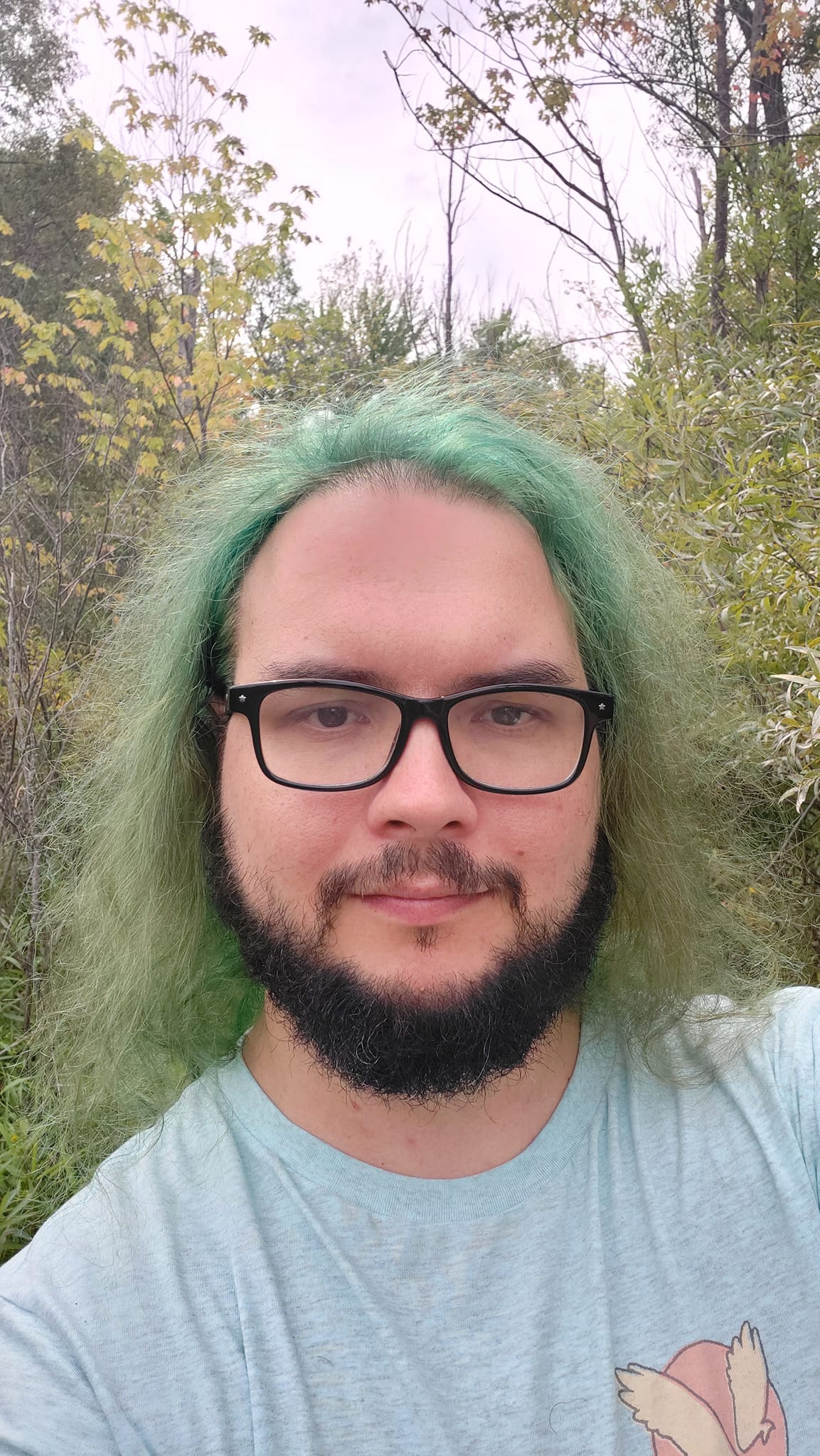 A man with green hair stands in front of marshside forest